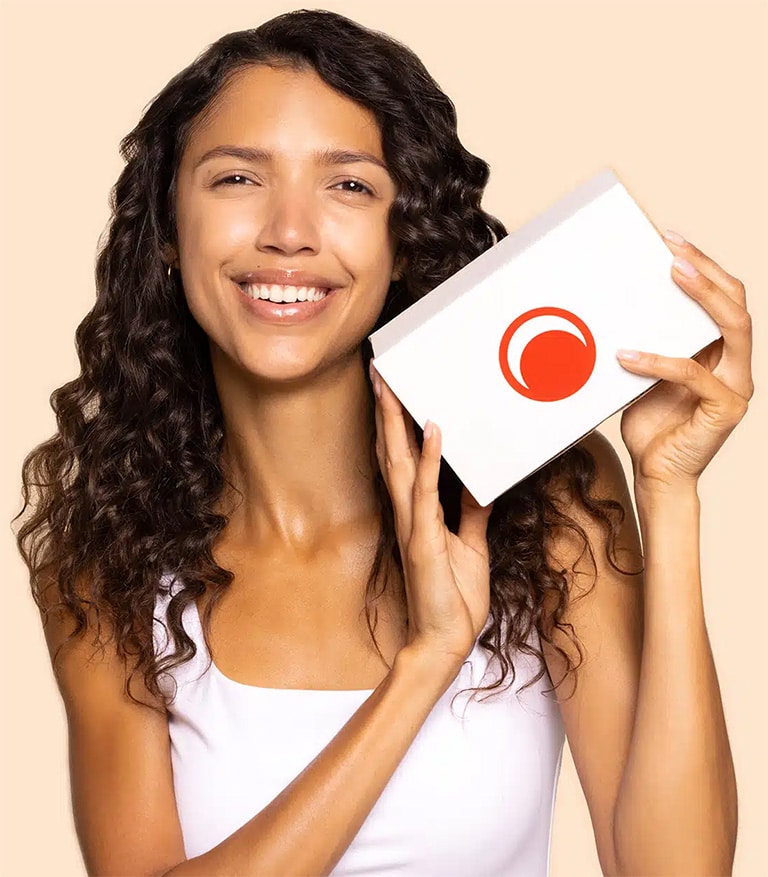 A woman with long curly brown hair, holding the Beauty Heroes subscription box, smiling